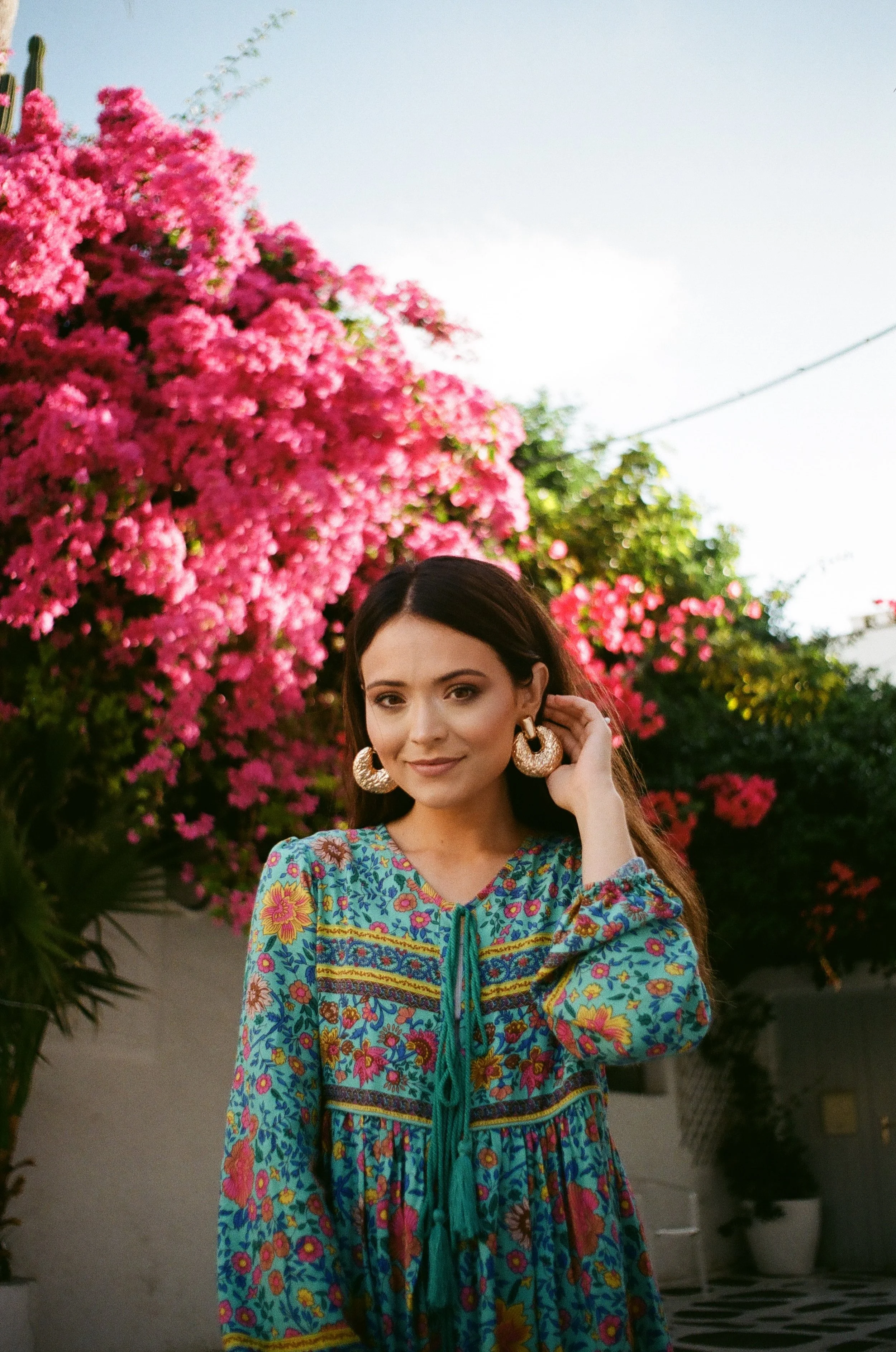 A woman wearing a colorful floral dress and large gold earrings standing outdoors in front of pink flowering bushes.