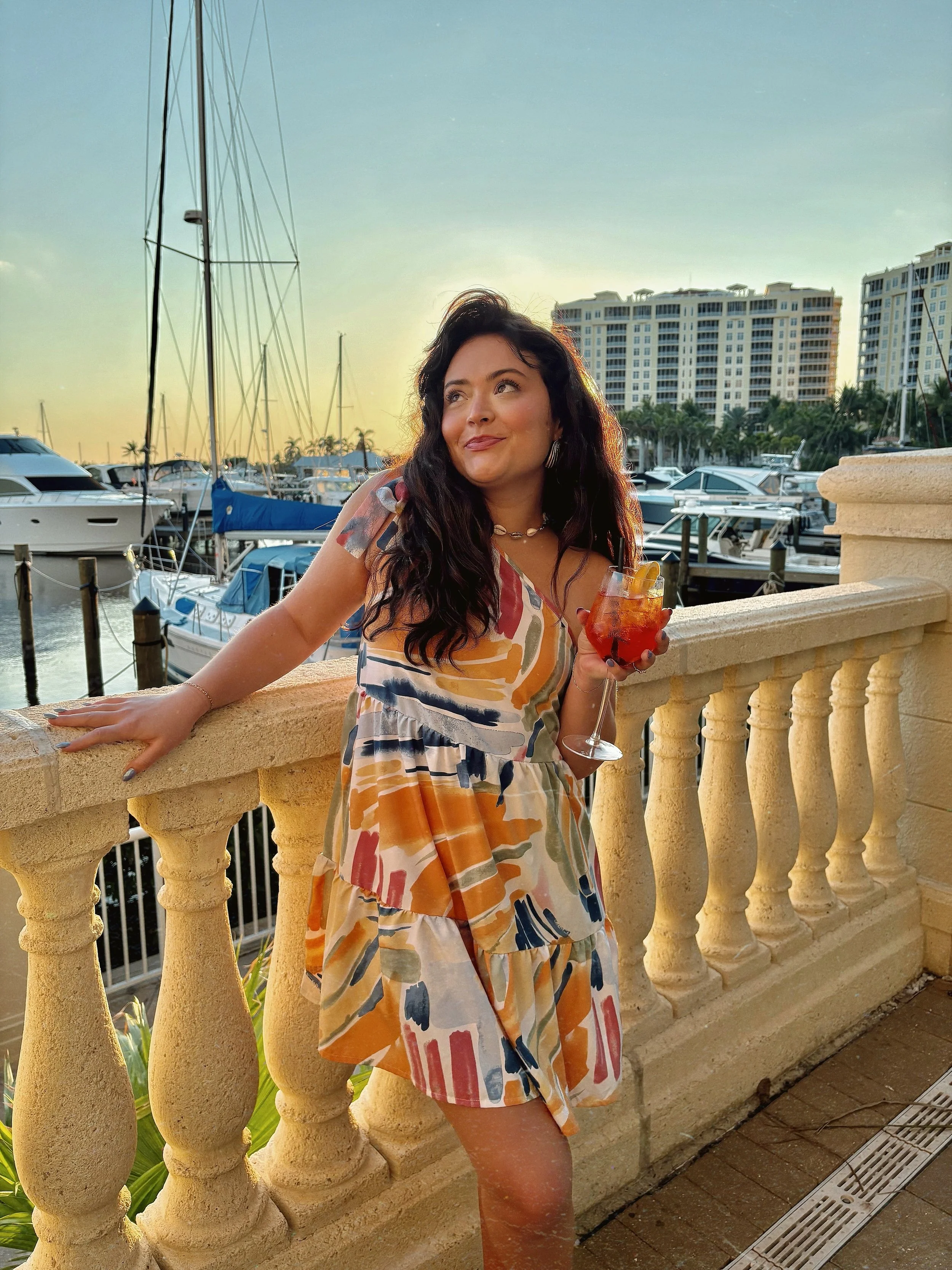 A woman stands on a balcony at sunset, holding a drink, with a marina full of yachts and high-rise buildings in the background.