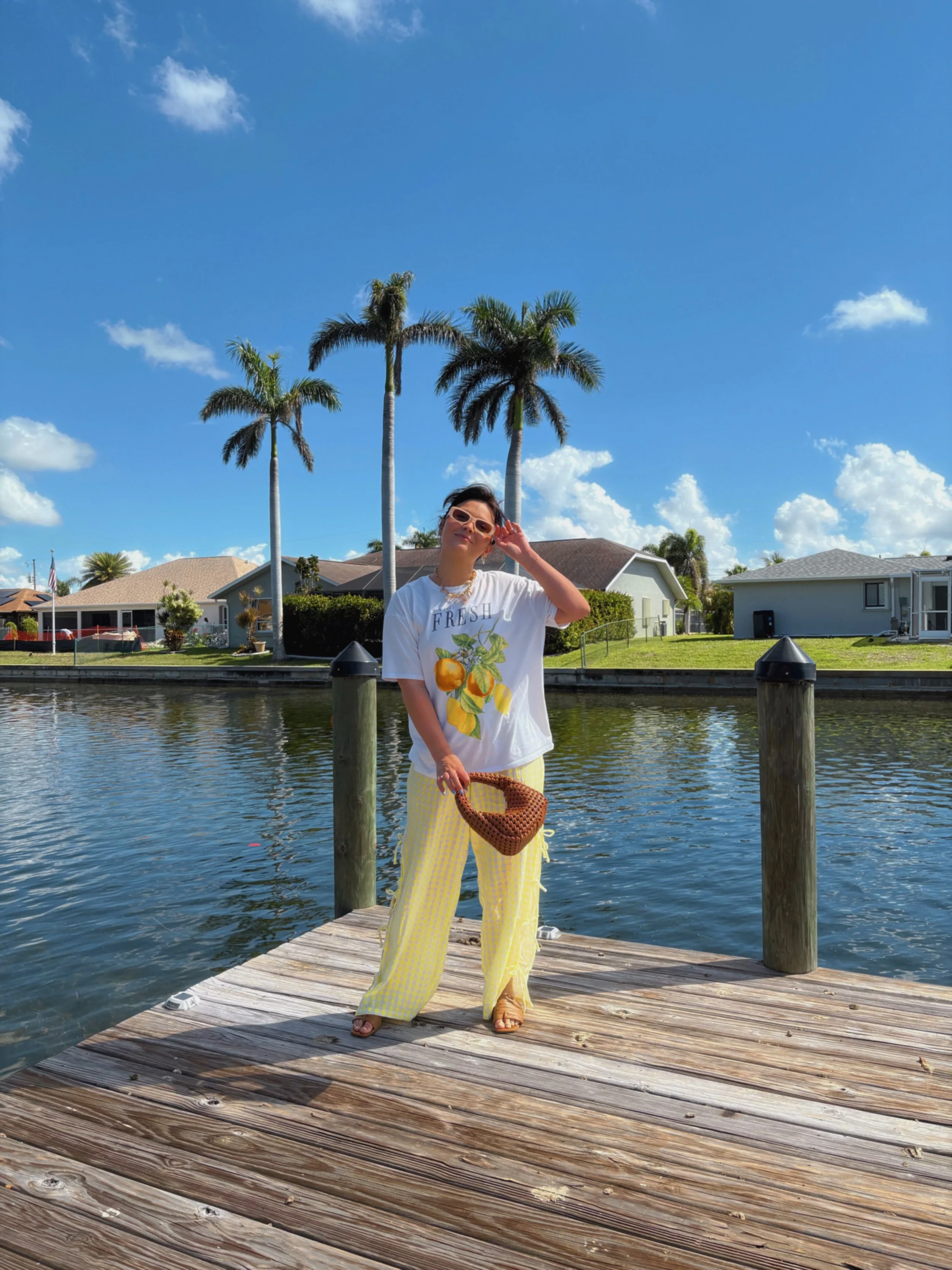 A woman standing on a wooden dock beside a canal, with palm trees and houses in the background, wearing a white t-shirt with lemon graphics, yellow striped pants, sunglasses, and holding a woven handbag.