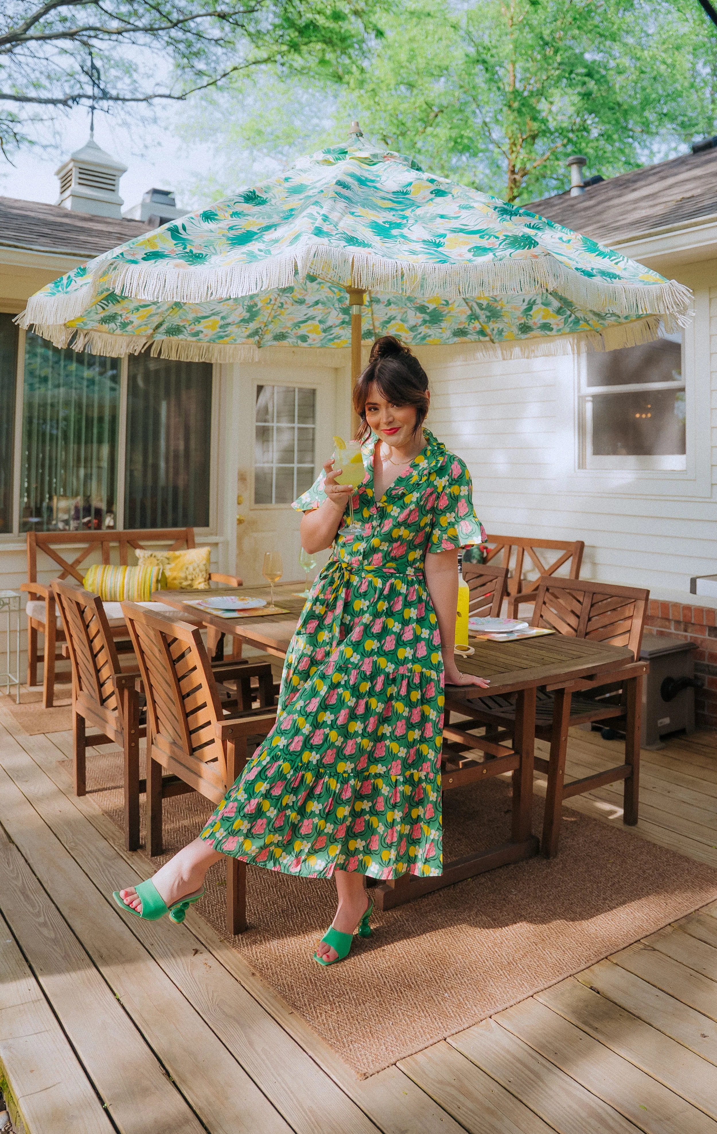 Woman in a green floral dress and green heels drinks a cocktail on a backyard patio with a wooden table, chairs, a large floral umbrella, and a white house with windows in the background.