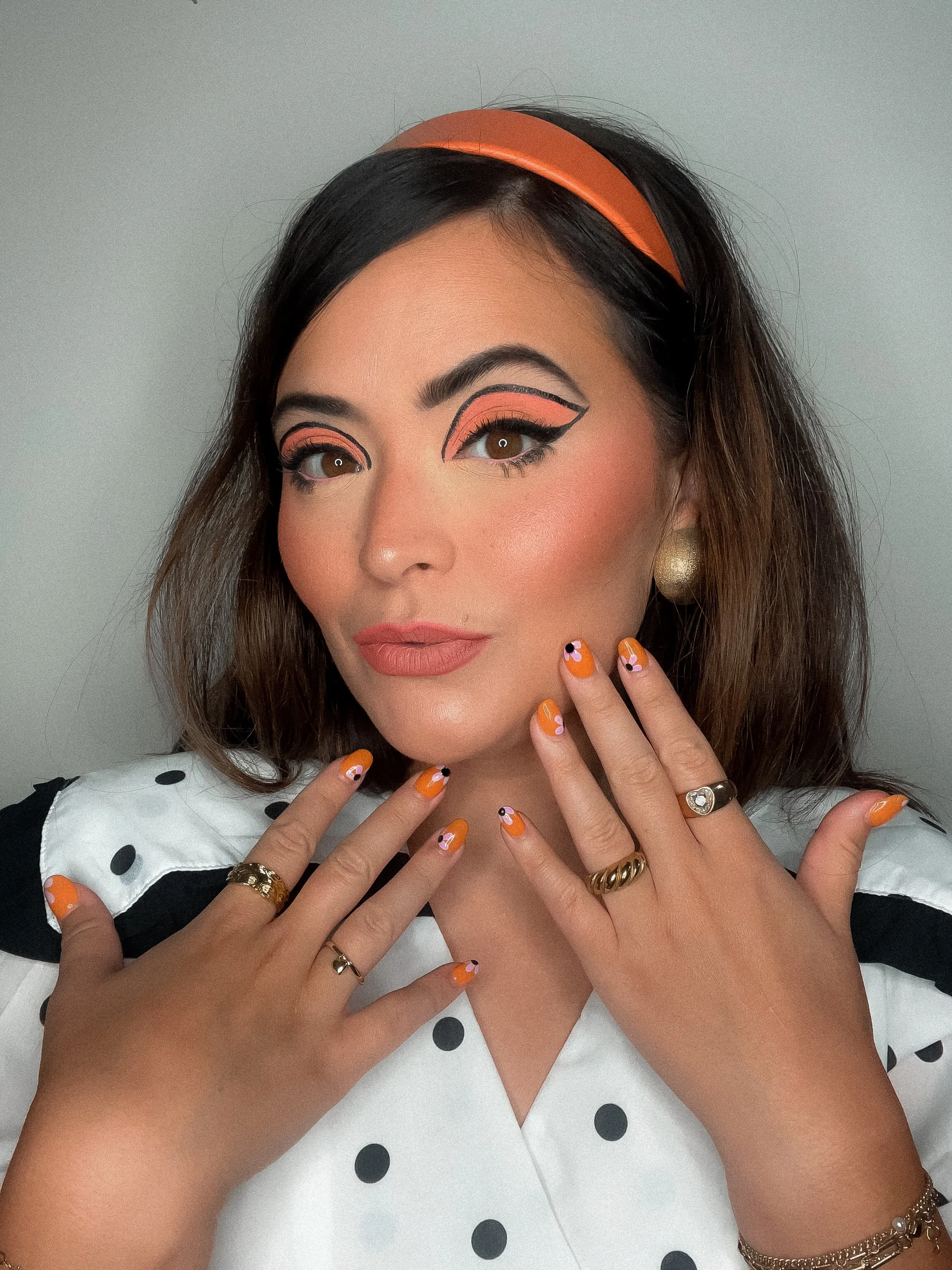 A woman with vintage-inspired makeup and polka dot clothing, wearing orange accents on her nails and headband, posing with her hands near her face.