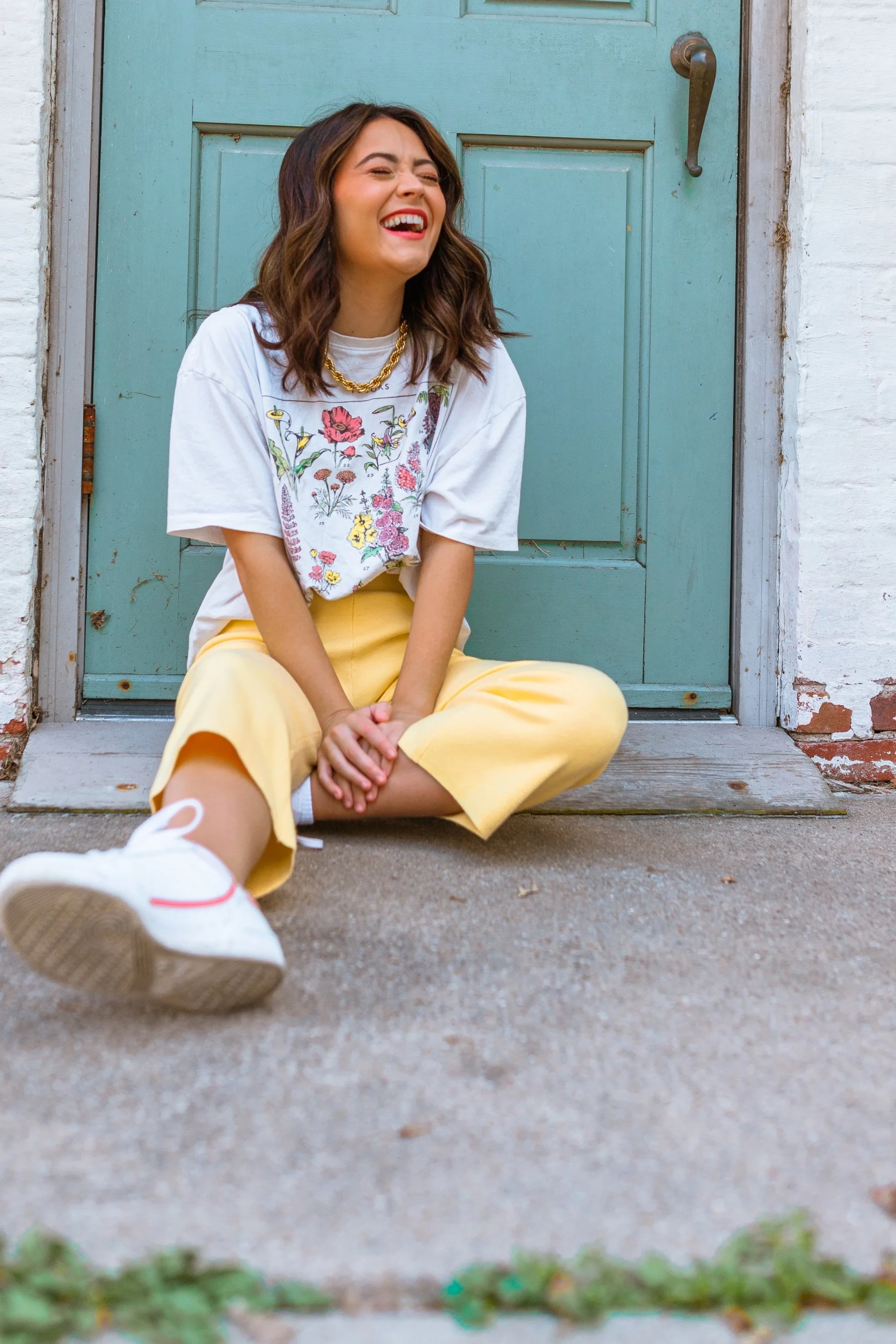 A young woman sitting on the ground in front of a blue door, laughing with her eyes closed, wearing a white embroidered T-shirt, yellow pants, and white sneakers.