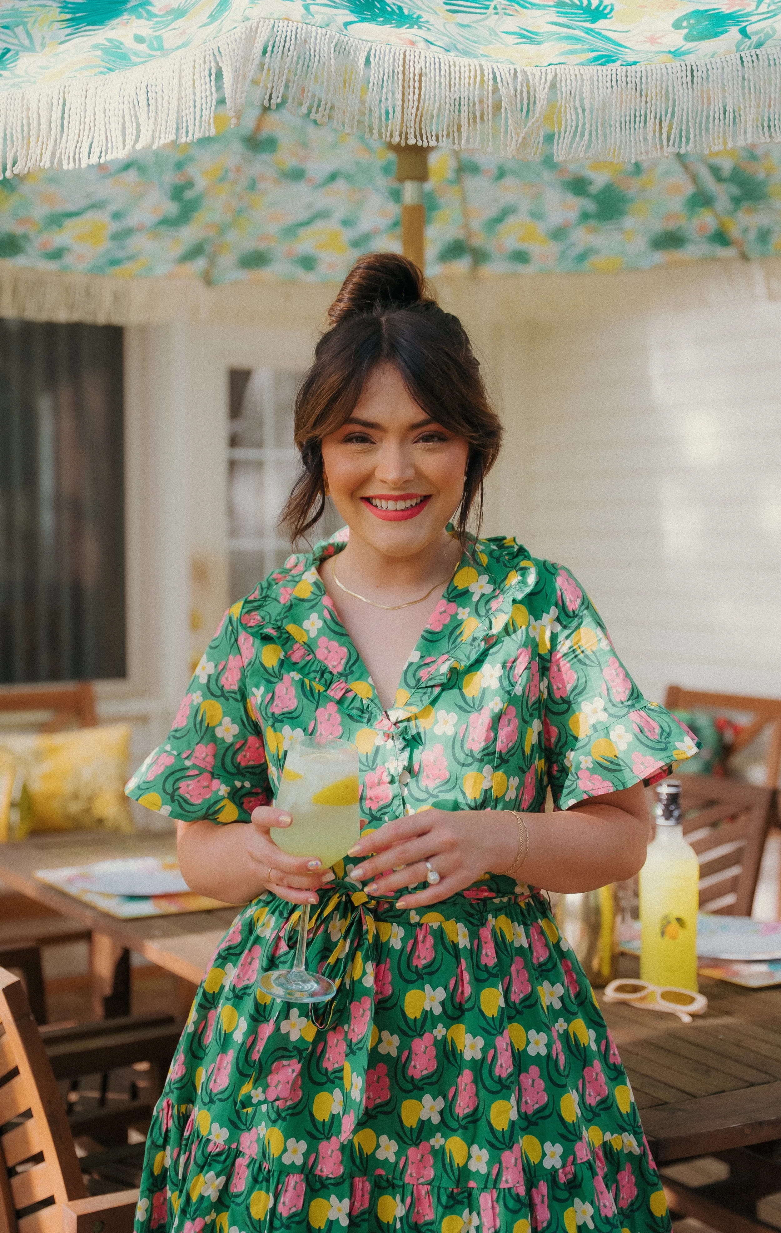 Young woman standing outdoors under a colorful umbrella, holding a glass of lemonade, smiling, wearing a floral dress.