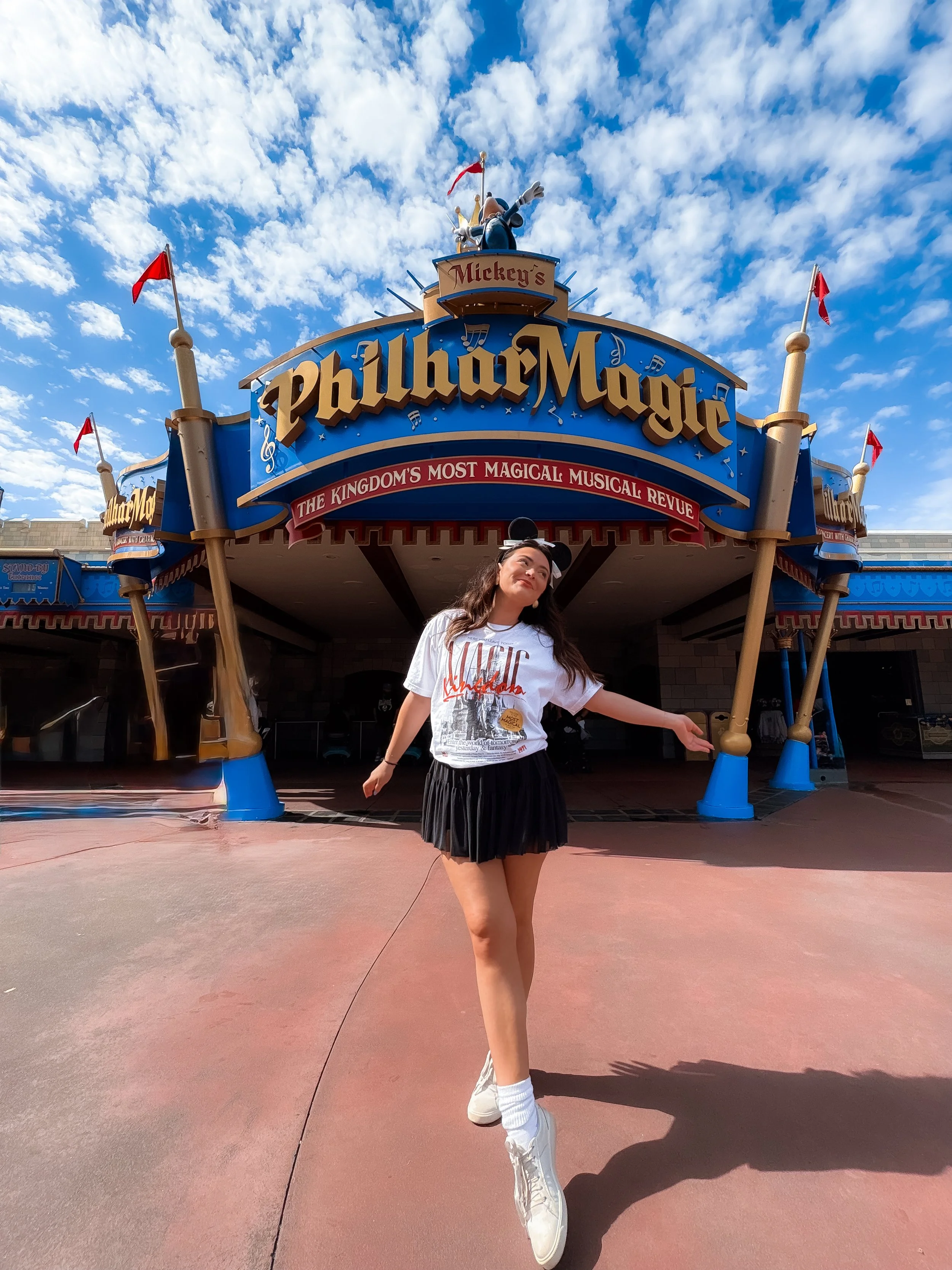 A young woman in a white t-shirt, black skirt, and sneakers posing in front of Mickey's Philharmagic at a Disney theme park on a sunny day with a partly cloudy sky.