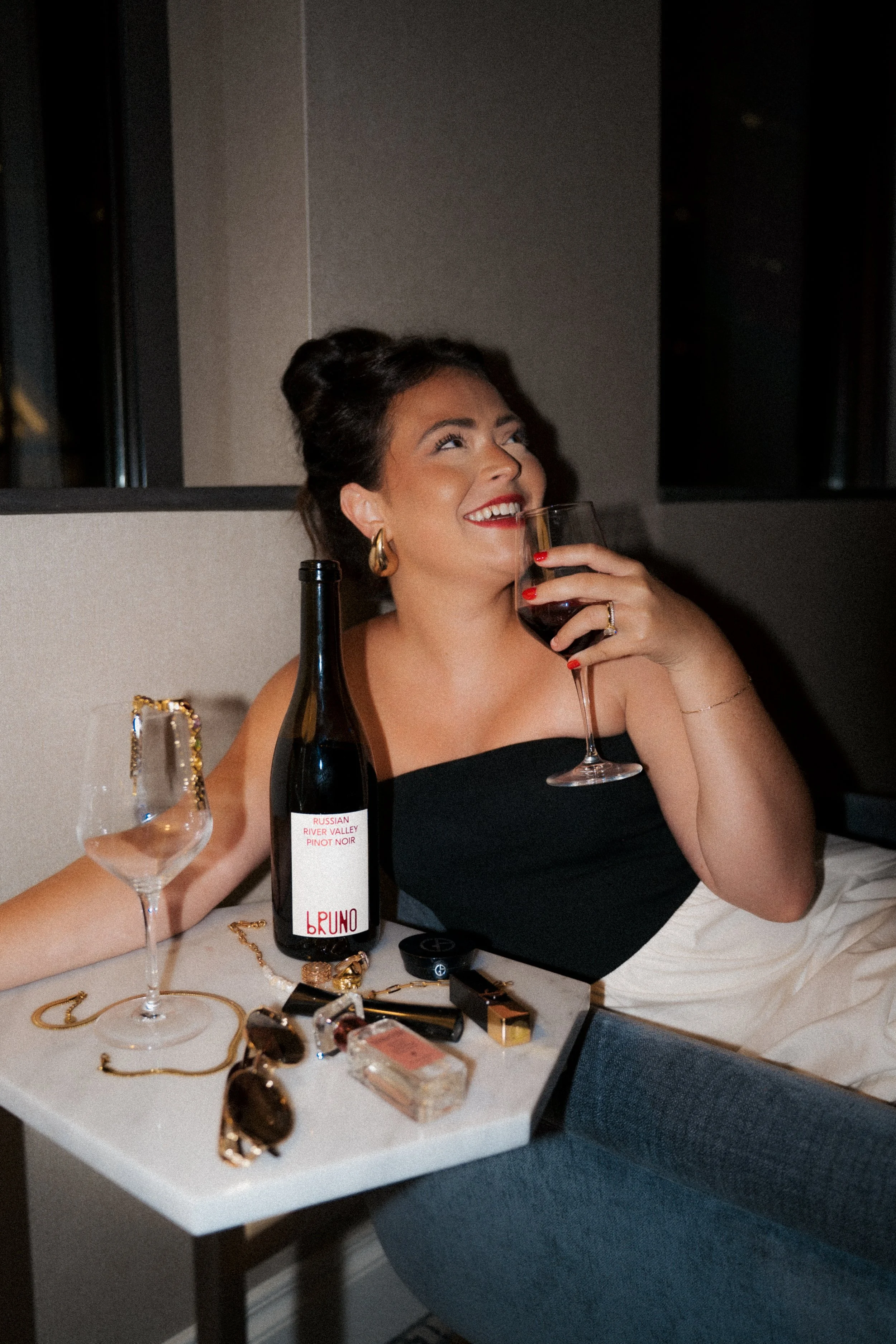 A woman with dark hair in an updo, wearing a black strapless dress, holding a glass of red wine, sitting at a table with jewelry, a bottle of wine, and sunglasses, smiling.