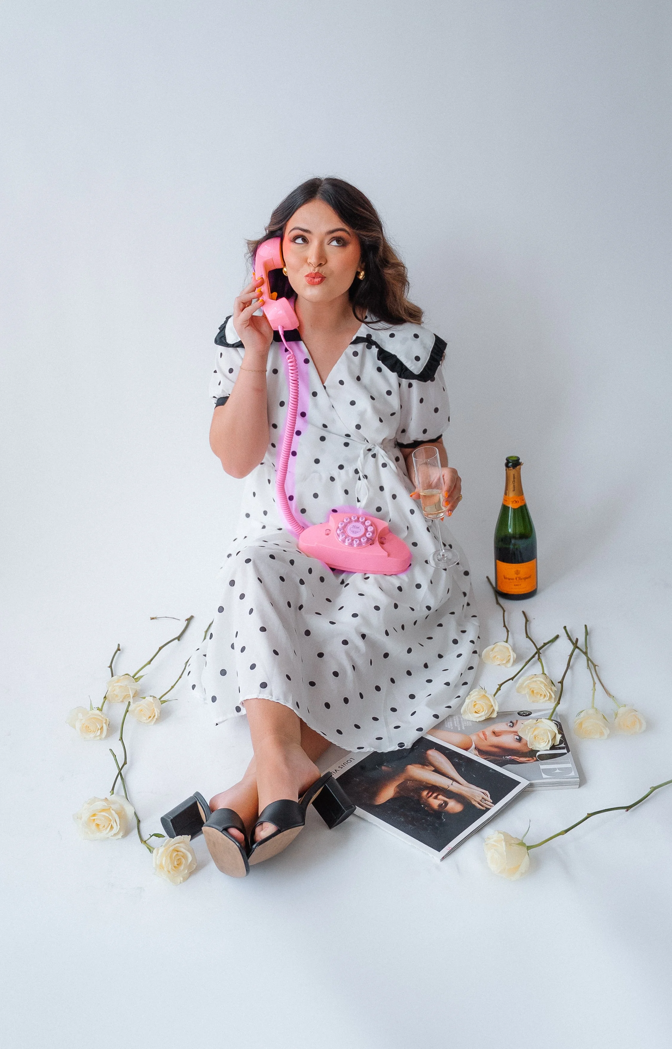 Woman in polka dot dress sitting on white floor, holding pink vintage telephone to her ear with a glass of champagne in hand, surrounded by white roses, a bottle of champagne, and magazines, with an open book in front of her.